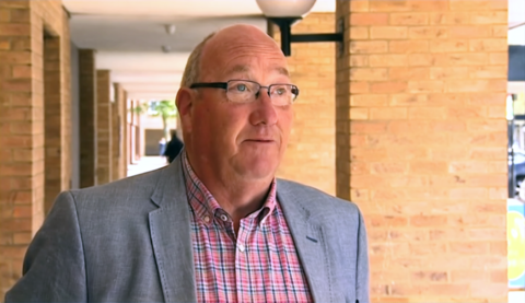 Robin Bradburn in light blue jacket with a red and blue checked shirt standing in the exterior corridor of a housing block. He is wearing glasses.