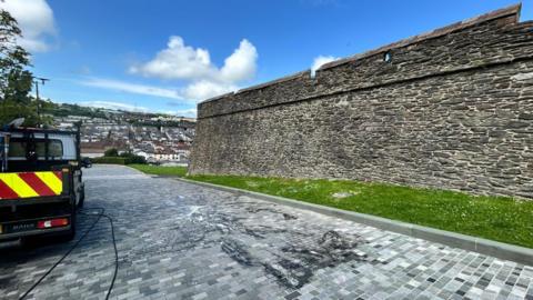 A dark-coloured stone wall with grass surrounding it. There is a grey brick road and in the distance is housing.  A van is parked with a hose to clean up the white splashes on the ground.