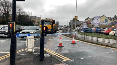 Traffic cones and police tape across a pedestrian crossing, with traffic queuing ahead.