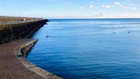 A view of St Catherine's breakwater. The sea, to the right, is blue and there are buoys dotted around. 