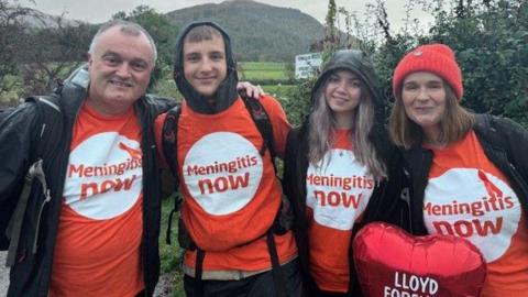 Tony Sutton, AJ Pearson, Ella Dixon and Louise Sutton smile at the camera before the start of the walk. They are wearing orange t-shirts reading Meningitis Now and black jackets. Louise, is also wearing a read beanie and holding a red heart-shaped balloon with Lloyd Forever written on it in white.  Ella and AJ have black hoods on. All four are wearing backpacks. 