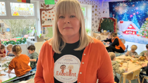 Lisa Owen, wearing a Chuckles nursery T-shirt, showing its "excellent" rating, in the nursery while children work at tables behind, with Christmas pictures on the wall