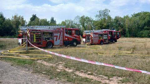 Fire engines are on a field that is cordoned off with red and white tape and long yellow hose reel jets are lying on the ground. There are trees around the edge of the field. The grass is yellow and dry.