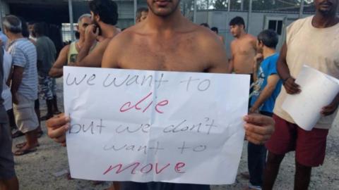 A man in a detention camp holds a sign reading, "We want to die, but we don't want to move".