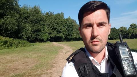 Handout photo issued by the Crown Prosecution Service of Ryan Connolly who is wearing a police uniform of white shirt and a black vest which has a communication device attached to it. He has straight dark hair, brushed into a side parting and has slight facial stubble. He looks directly into the camera, behind him a wide open space of grass is visible, with a path worn into it. Beyond that are trees in leaf and a bright blue sky