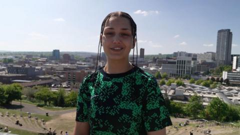 AJ is stood proud in front of the Sheffield landscape. She is wearing a black and green pattered T-shirt and her hair is in braids.