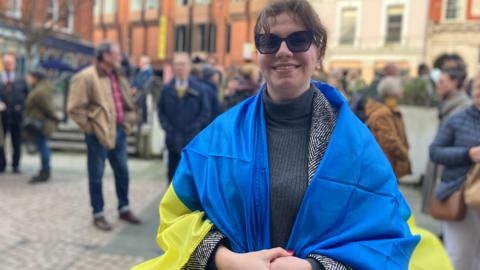 A woman with a blue and yellow flag round her shoulders stands in a market square with people behind her.