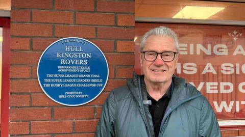 A man with white hair, he has a grey goat on and is smiling at the camera wearing glasses. He's stood outside of Craven Park's stadium next to the new blue plaque marking Hull KR's achievements. 