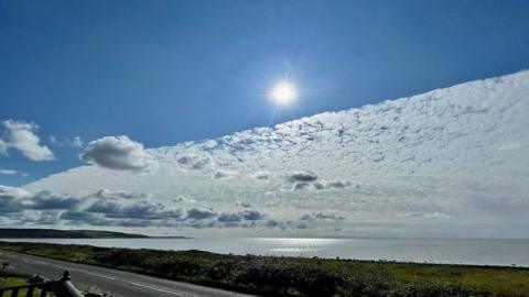 The sky, with the sun in the centre, is cut in half by a diagonal line with a cloud on one side and bright blue on the other. There is a bay with a road and grass verges to the front.