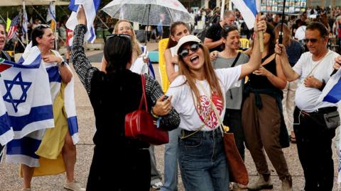 A group of people, with two women in front, linkinng hands and dancing, surrounded by others displaying Israeli flags, some with yellow umbrellas showing with 'NOW' printed on them