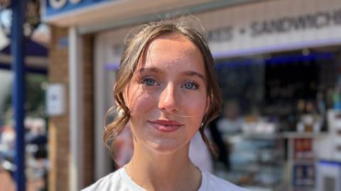 A head and shoulders photograph of 15-year-old Maisy. She has brown hair in a ponytail and is wearing a white T-shirt.