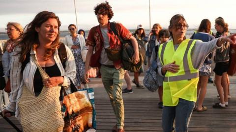 A woman in a striped shirt wheels her luggage. A volunteer wearing a high-vis vest is giving directions as other people carrying bags walk and chat.