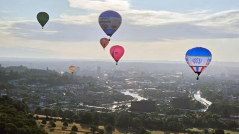 Hot air balloons flying over Bristol. The city and its harbour can be seen below, while the sun rises over the landscape.