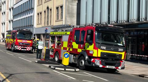 Two fire engines are parked next to buildings lining a road. There are a couple of firemen in uniform standing next to one of the engines. The engines are branded with red and fluorescent yellow checks.