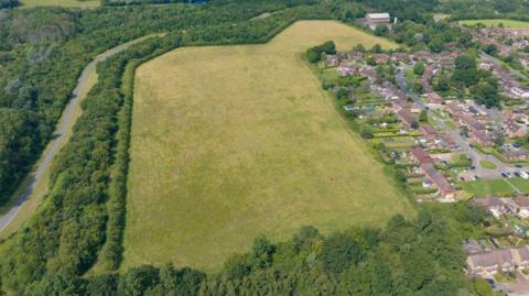 Grassland a developer wants to build on. It is surrounds by trees on three sides and buildings on one
