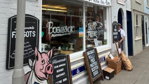 A butchers shop front has boards in front of the windows standing on the pavement advertising homemade meat products. A man wearing a white shirt and dark apron can be seen standing just inside the doorway.