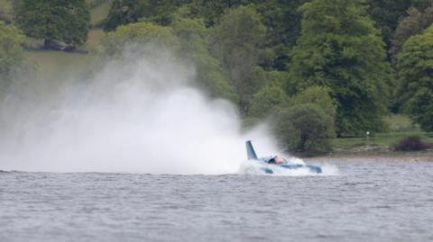 Bluebird - a craft that looks like a cross between a plane and a boat - leaves a plume of spray behind as it moves across Coniston Water. Trees fill the background.