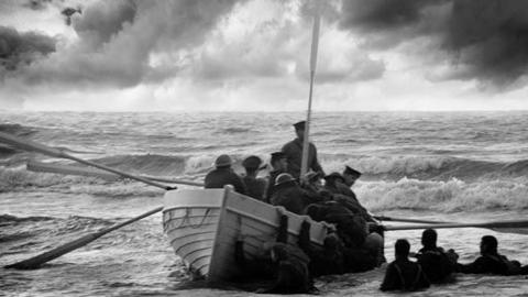 Black and white image of a lifeboat, oars in the water hanging over the side, with around 10 people onboard helping people in from the water.  Some of those in the water can be seen holding on to the side of the boat.  Three are seen treading water to the right hand side.