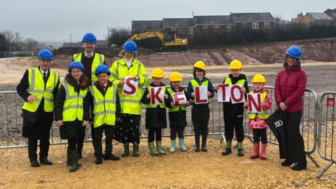 About a dozen people are standing in front of a grey metal fence on the construction site. Most of them are children and they are wearing high-vis jackets and hard hats. Some are holding up red letters that spell out: "Skelton". There is a yellow digger behind them and a row of red-bricked houses in the distance.