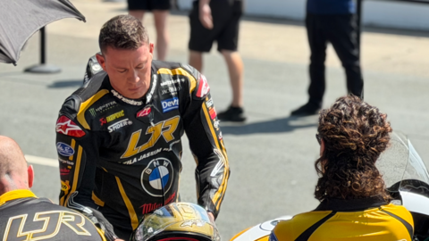 Channing Tatum is wearing black and gold leathers and is leaning over slightly, reaching for a matching helmet, with people stood around him, on a sunny day.
