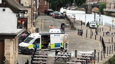 Picture shows a pub on the left with wooden tables and seats on a cobbled area outside the pub. Behind the tables and seat is a police van with it's side door open and towards the top of the picture is the river with another police car in the background.