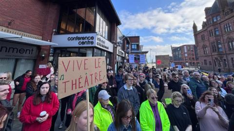 Shot of dozens of people standing outside in the centre of a market town on a sunny day. In the background is a building which says Conference centre - its a grand old brown brick building. One of the people has a cardboard sign which says Vive Le Theatre.