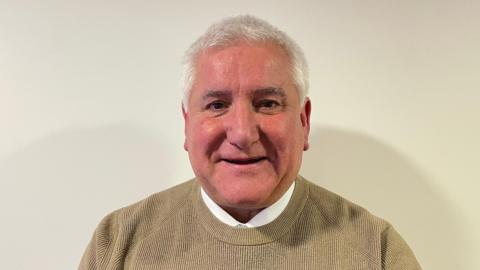 Councillor Patrick Harley. He has short white hair, brown eyes and is clean shaven. He wearing a brown jumper and is stood in front of an off-white background. 
