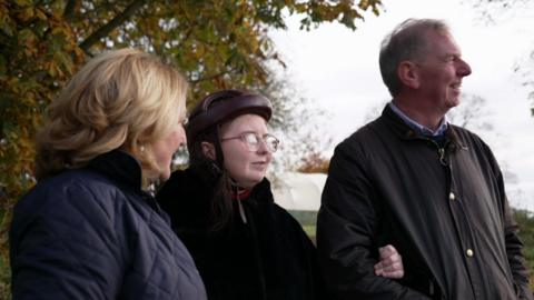 Emily in centre has brown hair and is wearing glasses and a black coat, with her parents. Her mum has blonde hair and is wearing a blue jacket, her dad is has short hair and is wearing a brown jacket.