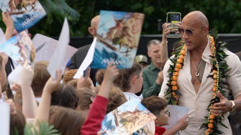 A man with no hair is wearing a button up shirt and sunglasses, speaking to school children holding posters in the air