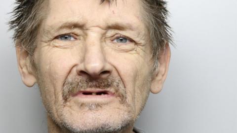 A custody picture of a man with messy grey hair and facial stubble. 