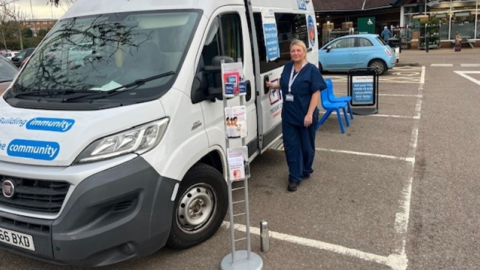 A member of nursing staff stands outside the minibus in navy uniform. The grey vehicle is parked in a car park and there is a stand containing leaflets near its front.
