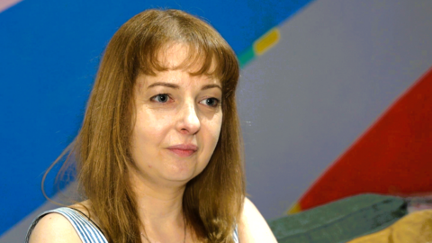 A woman with brown hair looking off to the right of the screen. She is sat on a sofa. Behind her is a white, red and blue wall.