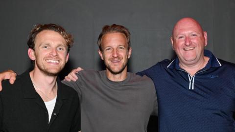 Three men stand in a line with arms around each other and smile at the camera. Two are in their thirties or forties with dark blond hair. One wears a black shirt, the other a grey t-shirt. The third man is older, bald and wears a navy polo shirt.