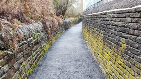 A freshly-laid blue-black tarmac footpath with stone walls either side coverage in yellow and green moss.