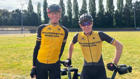 Two men wearing mainly yellow tops including the words Wolverhampton Wheelers. They are wearing glasses and cycling helmets and a large area of grass and trees are in the background.