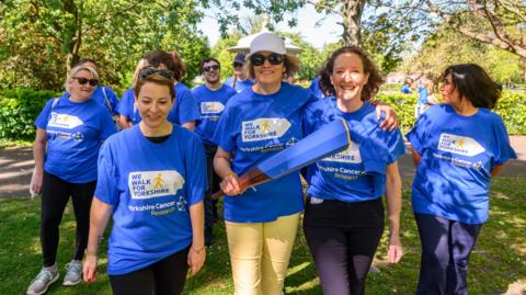 A group of women in blue t-shirts walk together through a park. One is holding a baton which looks liek a cricket bat and is painted blue. 