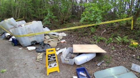 A roadside area in a wooded setting with dumped rubbish, including plastic containers, furniture pieces, and boards, alongside yellow tape and a warning sign.
