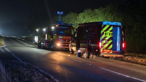 A fire engine and a smaller van from the fire service, parked on the side of a road, next to a hedge. It is dark and both vehicles have their beam lights on. There are bags on the floor, next to the van.