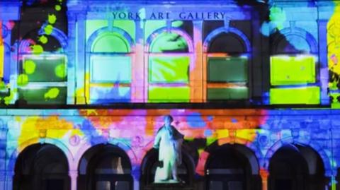 A Victorian art gallery building with a  series of open arches and a statue in front with multi-colour images projected onto the building