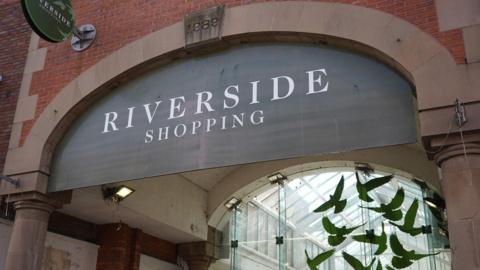 A black sign which says "Riverside Shopping" in white letters over the entrance to a shopping centre 