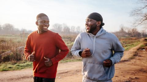 Two young men jog on a dirt path in a park, smiling to the other. One is wearing a red jumper, the other is in a grey jumper.