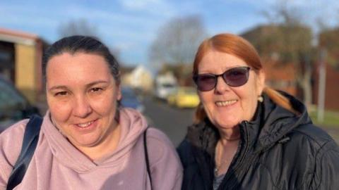 Two women are standing in a car park. The woman on the left is wearing a light pink hoodie with a rucksack on. The woman on the right is wearing a black padded coat with sun glasses. Both women have their hair tied back. 