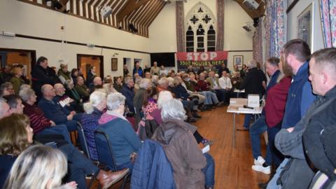 A large crowd of people in a village hall listening to someone speak. There is a banner on the far wall reading "Stop 3500 houses here"