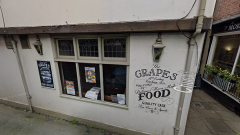 The outside of the Grapes Tavern. It is a white building with the name printed on the side in black lettering. The window contains posters for events, and there is an advert for burgers to the left of the window.