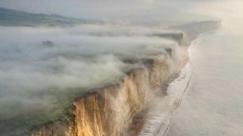 Thick fog rolls off the top of cliffs and into the sea. The sky is grey and foggy. The cliffs have a beige colour to them. A calm sea is rolling onto the shore at the foot of the cliffs.