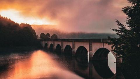 A photo of a bridge over Ladybower Reservoir in the Peak District as the early morning mist began to lift. The light is low and reflecting on the water, and it is cloudy. 