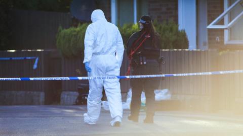 Forensic officer in white suit and police woman in dark uniform with their backs to camera, stood outside a row of bungalows with a low wooden fence, there is police tape cordoning off the road