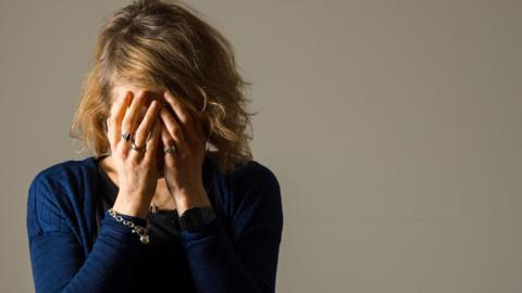 A woman is holding her face in her hands. Her wavy blonde hair reaches her shoulders. She is wearing a blue jumper and has rings, a necklace and a bracelet.
