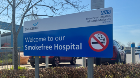 A mainly blue and white sign by a hedge includes the words "Welcome to our Smokefree Hospital" and a red, white and black no-smoking sign. Some cars are visible in the background.