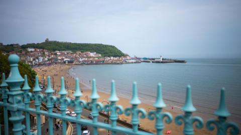 Blue railings in the foreground. Behind them there is a view of Scarborough South Bay which shows the curve of the beach and part of a road running along the seafront.